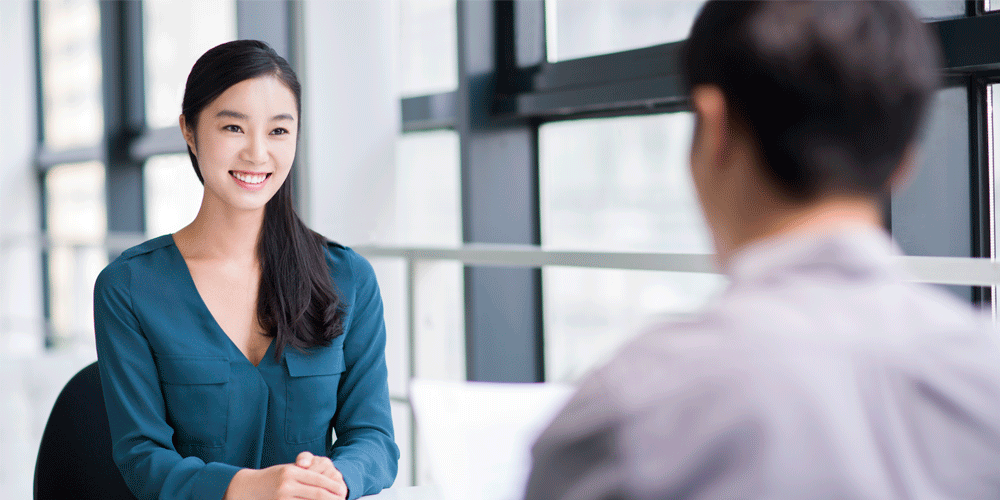A young woman sitting at a table across from someone out of frame who interviews her.
